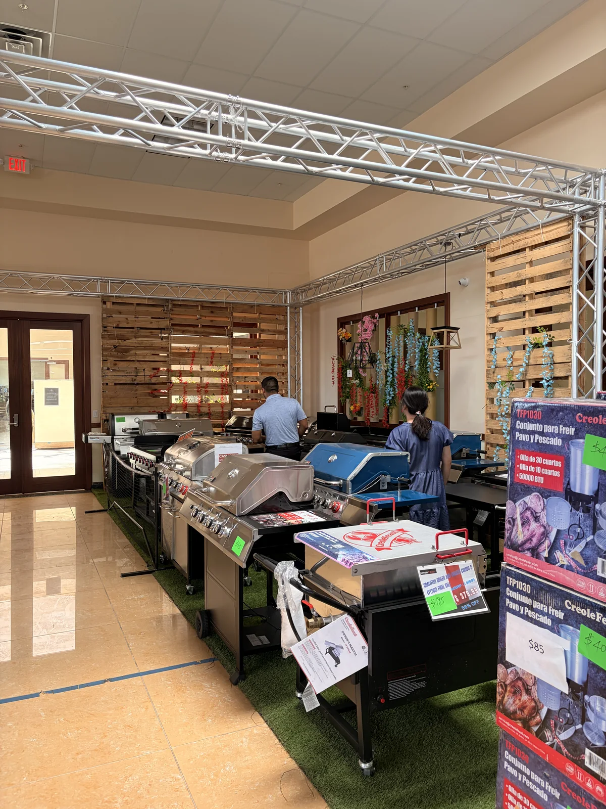 Gas grills lined up on turf with handwritten price tags at the Cube-Age Houston pop-up sale