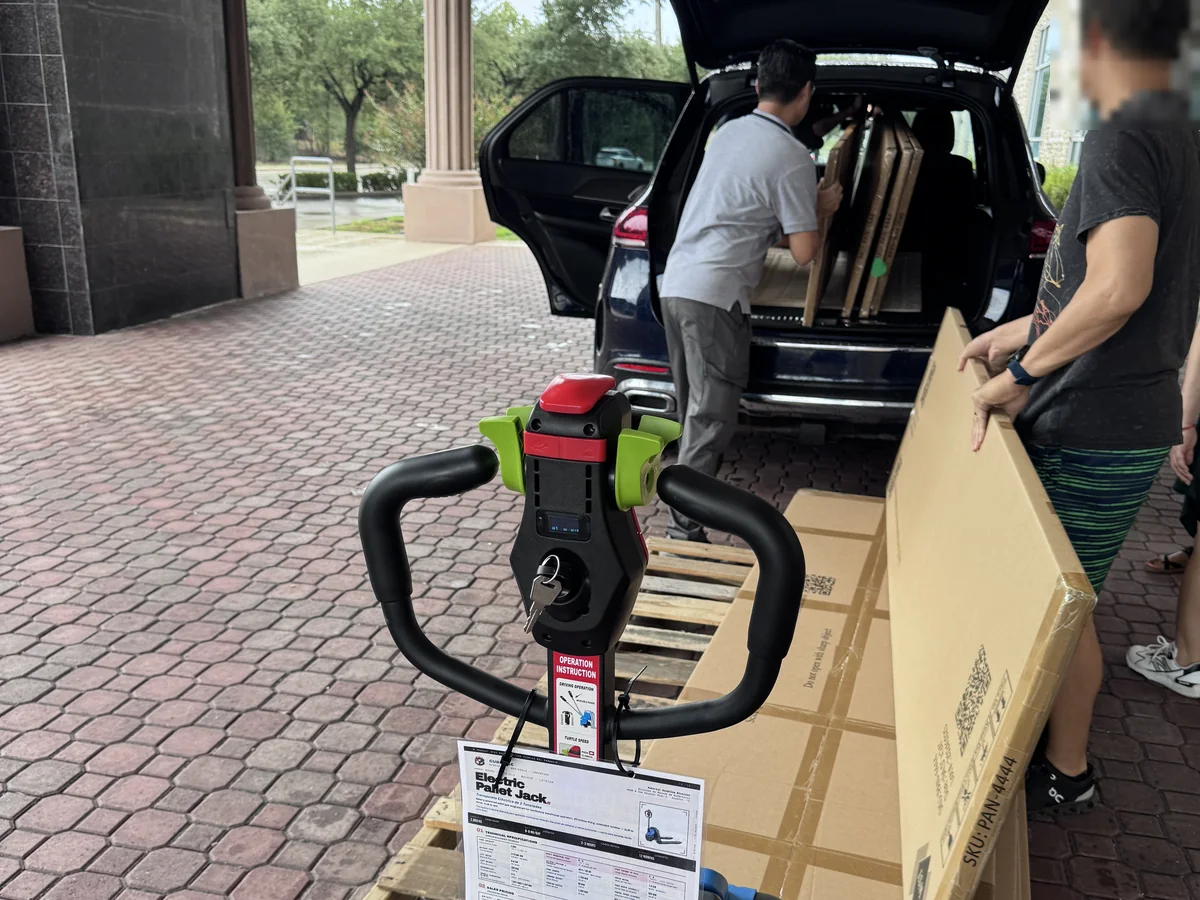 A customer loading a pallet of boxed wall panels into an SUV, with an electric pallet jack in front, at the Cube-Age Houston sale