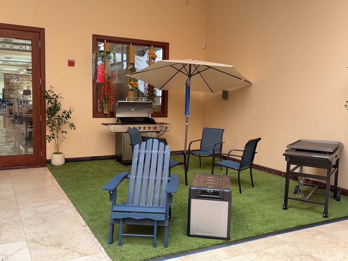 The finished outdoor scene at the showroom entrance: patio chairs, a gas grill, a griddle and a parasol on turf