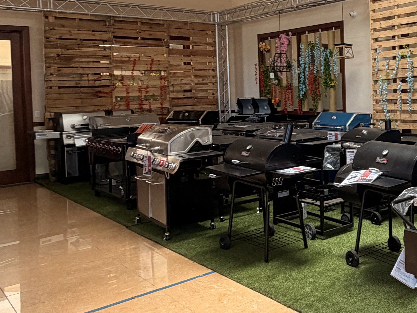 Gas grills and flat-top griddles lined up on turf against the pallet wall in the Cube-Age showroom
